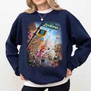 Può includere: Felpa blu navy con una grafica colorata del film d'animazione "Zootopia 2". Il design include vari personaggi animali e il titolo del film su un cartello stradale. La felpa è a girocollo.