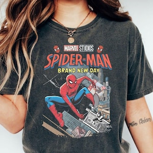 Op de afbeelding: Donkergrijs t-shirt met een Marvel Studios Spider-Man graphic. Het ontwerp bevat de tekst "Spider-Man Brand New Day" en een illustratie van Spider-Man in actie tegen een stadsgezicht. Het shirt heeft een ronde hals en korte mouwen.