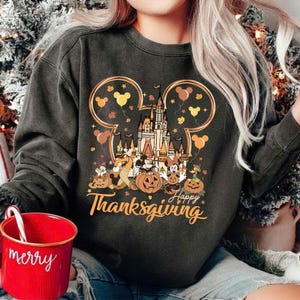 Op de afbeelding: Donkergrijze sweatshirt met een kleurrijke afbeelding met een kasteel, Mickey Mouse, pompoenen en de woorden "Happy Thanksgiving". Het ontwerp is omlijst door Mickey Mouse oren en ballonnen. Een rode mok met "merry" staat op de voorgrond.