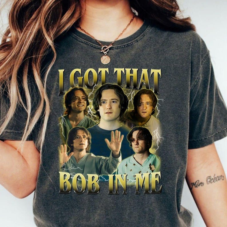 May include: Dark gray t-shirt with the text "I GOT THAT BOB IN ME" in gold lettering. The shirt features a collage of images of a person with long brown hair, with lightning bolt graphics.