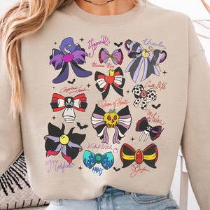 May include: Beige sweatshirt featuring colorful bow designs inspired by Disney villains. Each bow is unique, with names like "Ursula" and "Maleficent" written above them. The bows are in various colors, including purple, red, and black.
