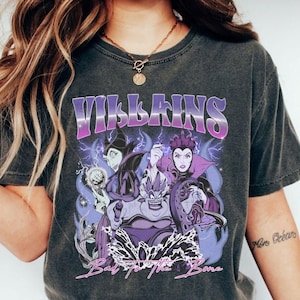 May include: Dark gray t-shirt featuring the word "VILLAINS" in a purple and white gradient font. The graphic includes illustrations of cartoon villains with purple and blue accents, and the text "Bad to the Bone".