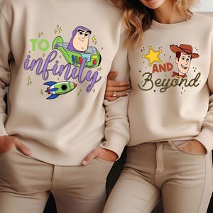 May include: Beige sweatshirts with cartoon graphics. One features Buzz Lightyear with the text "To Infinity", and a rocket ship. The other has Woody with a sheriff badge and the text "And Beyond".