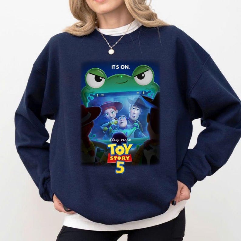 May include: Navy blue sweatshirt featuring a graphic from the Disney Pixar movie Toy Story 5. The graphic includes the characters Woody, Buzz Lightyear, and Jessie, with the text "IT'S ON." and "Toy Story 5".