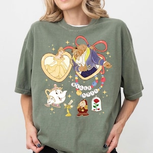 以下が含まれることがあります： オリーブグリーンのTシャツ。美女と野獣のキャラクターのカラフルなグラフィックが特徴です。デザインには、ベル、野獣、ミセス・ポット、チップ、ルミエール、コグスワース、ガラスケースに入ったバラが含まれています。「BELLE」という単語がビーズで綴られています。