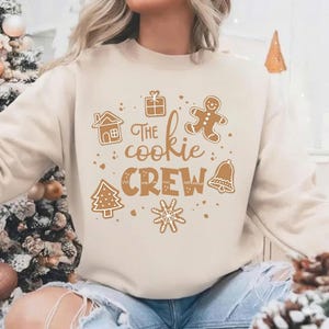 May include: A cream-colored sweatshirt with the text "The Cookie Crew" in brown, along with gingerbread-themed illustrations. The design includes a gingerbread house, gift, gingerbread man, Christmas tree, bell, and snowflake.