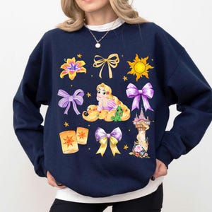 Op de afbeelding: Marineblauwe sweatshirt met een kleurrijke afbeelding van een prinses met lang blond haar, omringd door strikken, bloemen, een zon, een kasteel en lantaarns. Het ontwerp is een speelse illustratie.