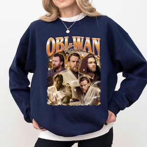 May include: Navy blue sweatshirt featuring a graphic of Obi-Wan Kenobi in various poses, with the name "Obi-Wan Kenobi" in an orange and gold retro font. The design includes lightning bolt accents.