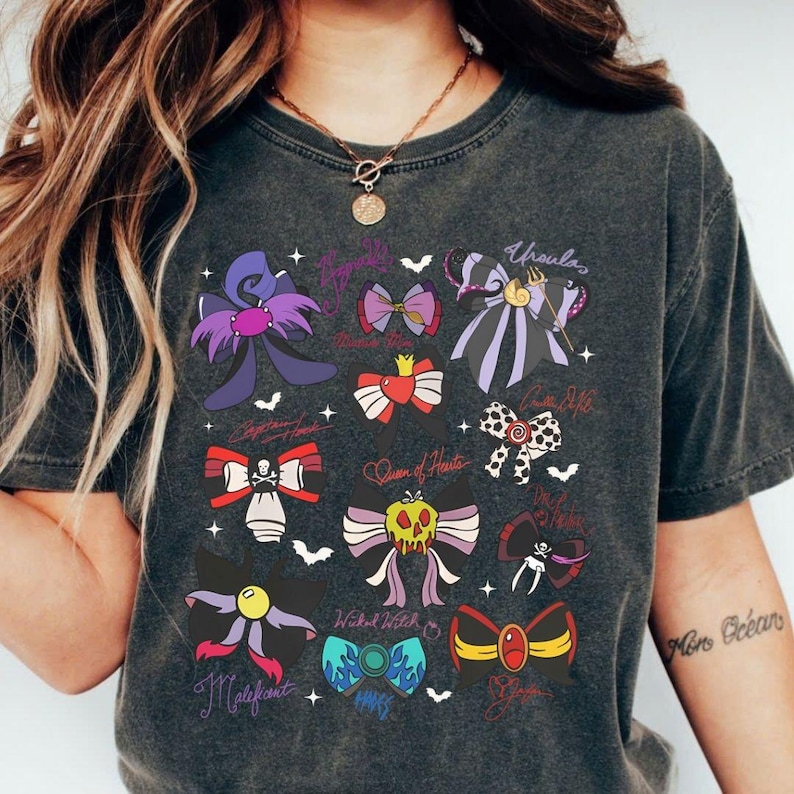 May include: Dark gray t-shirt featuring colorful bow illustrations with character names like Ursula and Maleficent. The bows are in various colors, including purple, red, and yellow, with a few small bat graphics.