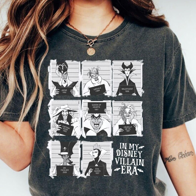 May include: Dark gray t-shirt featuring a grid of Disney villains, each holding a sign with their name and year. The shirt includes Jafar, Ursula, Maleficent, Captain Hook, Queen of Hearts, Cruella de Vil, Dr. Facilier, and Evil Queen. Text on the shirt reads "IN MY DISNEY VILLAIN ERA."