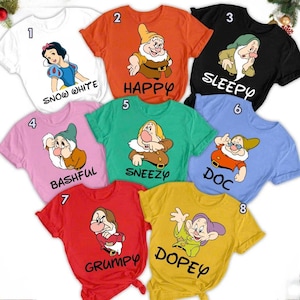 May include: Collection of eight colorful t-shirts featuring cartoon characters. Each shirt displays a different character from Snow White and the Seven Dwarfs, with the character's name printed below. Colors include white, orange, black, pink, green, blue, red, and yellow.