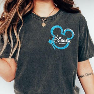 May include: Dark grey t-shirt with a blue glitter Mickey Mouse design, the words "Disney Channel" and "And You're Matching". The design includes a blue magic wand and stars.
