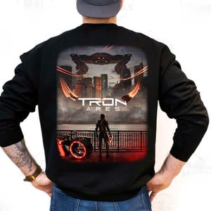 Puede incluir: Sudadera negra con un gráfico de "Tron: Ares". El diseño muestra una ciudad futurista, una figura y un vehículo brillante, con el título de la película. La obra de arte está en tonos de rojo, negro y gris.