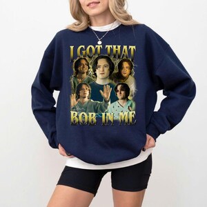 May include: Navy blue sweatshirt with the text "I GOT THAT BOB IN ME" in gold lettering. The graphic features a collage of images of a person. The sweatshirt is a crew neck style.