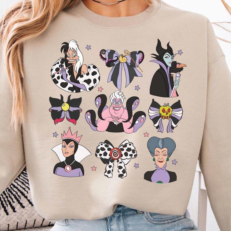 Op de afbeelding: Beige sweatshirt met kleurrijke illustraties van Disney-schurken, waaronder Cruella de Vil, Maleficent en Ursula. Het ontwerp bevat strikken en portretten van de personages, met steraccenten. De sweatshirt is een casual, comfortabele stijl.