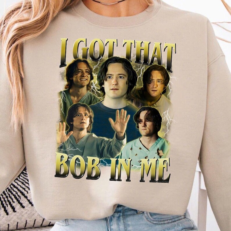 May include: Beige sweatshirt with a graphic featuring multiple images of a person with the text "I GOT THAT BOB IN ME" in a gold and black font. The design includes lightning bolt accents.