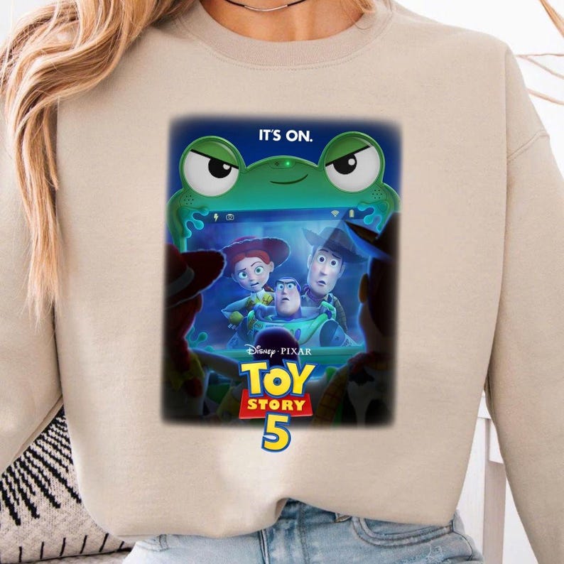 May include: A beige sweatshirt featuring a graphic from the Disney Pixar movie Toy Story 5. The graphic includes the text "IT'S ON." above a green frog-like character and the movie's title and number. The sweatshirt is a neutral color.