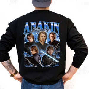 Op de afbeelding: Zwart sweatshirt met een afbeelding van Anakin Skywalker en de tekst "ANAKIN Skywalker" in het blauw. Het ontwerp bevat meerdere afbeeldingen van het personage met blauwe bliksem, in vintage stijl.