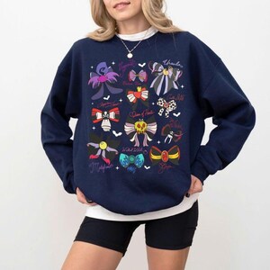 May include: Navy blue sweatshirt featuring a colorful bow design with character names. The bows are in various colors and styles, with names like Ursula, Maleficent, and Queen of Hearts. The sweatshirt has a crew neck and long sleeves.