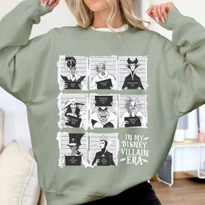 May include: Sage green sweatshirt featuring a grid of Disney villains in mugshot style, each holding a sign with their name and year. The text "IN MY DISNEY VILLAIN ERA" is printed below the grid in white.