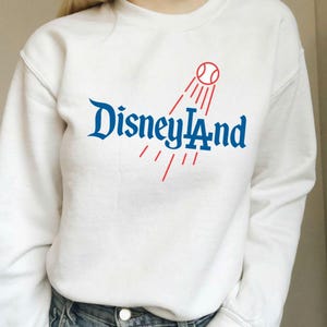 May include: White sweatshirt with the text "Disney LA" in blue script, with a red baseball and lines design. The sweatshirt is a casual style, suitable for everyday wear.