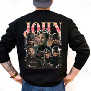 May include: Black sweatshirt featuring a graphic of John Walker with the name "John Walker" in a retro font. The design includes multiple images of the character in various poses. The sweatshirt has long sleeves and a crew neck.