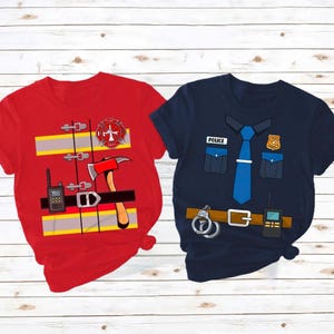 May include: Two short-sleeved t-shirts with costume designs. One is red with a firefighter theme, including a fire axe and radio. The other is navy blue with a police officer theme, featuring a tie and badge.