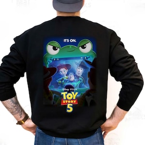May include: Black sweatshirt featuring a colorful graphic from the movie Toy Story 5. The graphic includes the text "IT'S ON." above a green frog and the Toy Story characters Woody, Buzz Lightyear, and Jessie. The Disney Pixar logo is also visible.