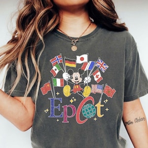 May include: Dark gray t-shirt featuring Mickey Mouse holding flags from various countries. The word "Epcot" is written in colorful letters below the character, with a globe graphic. The shirt has a vintage, distressed look.
