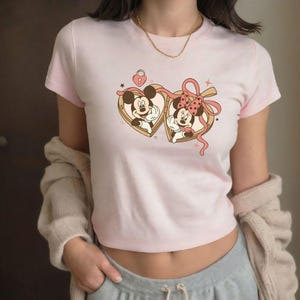 Op de afbeelding: Een lichtroze crop top met een afbeelding van Mickey en Minnie Mouse in hartvormige medaillons. Het ontwerp bevat een roze strik, sterren en een slot. De top heeft korte mouwen en een ronde hals.