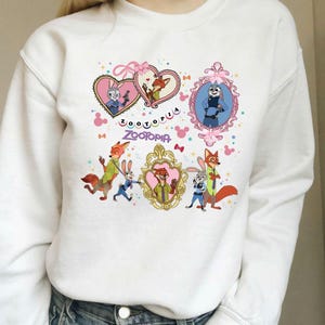 May include: White sweatshirt featuring colorful illustrations of characters from the animated film "Zootopia." The design includes framed portraits, hearts, and the film's title. The artwork is in shades of pink, blue, gold, and green.