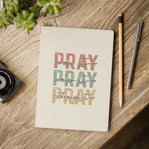 May include: A cream-colored notebook with the text "Pray on it, Pray over it, Pray through it" in a vintage style font with hearts. The text is in shades of red, green, and yellow.