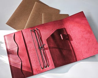 May include: Open red leather journal with pockets and a strap closure. The journal is open, revealing a dark red interior and black elastic cords. Several brown paper notebooks are visible in the background.