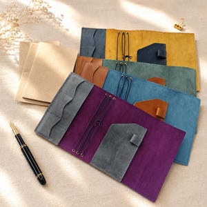 Puede incluir: Una colección de fundas rectangulares de cuero para cuadernos en morado, azul, verde y amarillo, cada una con bolsillos internos, encuadernación con cordón elástico y un lazo para bolígrafo. Incluye una pluma estilográfica negra y papel beige.