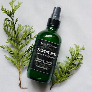 May include: A green glass bottle with a black label that reads "ROBES OF DREAMS FOREST MIST room & body spray refreshing + clearing pine cedar amyris rosemary ALL NATURAL".