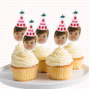 May include: Five cupcakes with white frosting and pink and green cupcake toppers with a child's face and the word "ONE" on each topper.