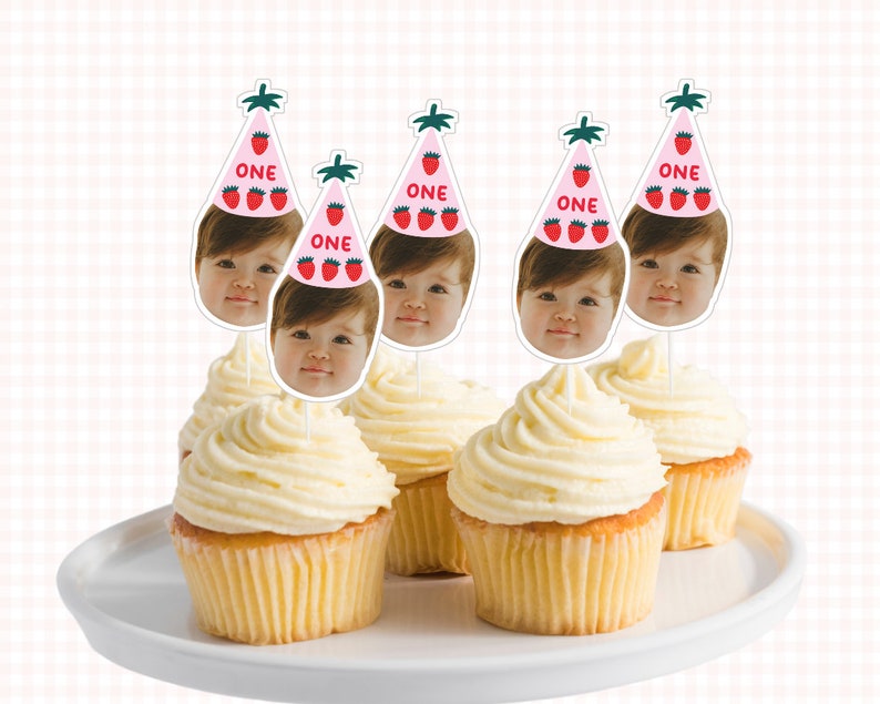 May include: Five cupcakes with white frosting and pink and green cupcake toppers with a child's face and the word "ONE" on each topper.