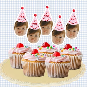 May include: Six pink cupcakes with white icing and colourful sprinkles, topped with a red cherry. Each cupcake has a pink and red party hat with the word "ONE" and a cherry design, and a baby's face peeking out from the top.