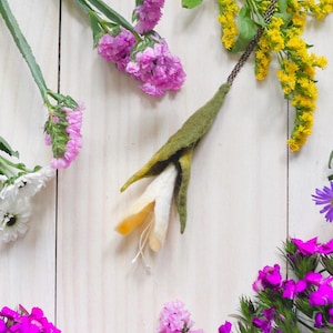 May include: A handmade felt flower pendant necklace with a green stem and a white and yellow flower. The necklace is surrounded by various colorful flowers, including purple, yellow, and white blooms, arranged on a light wooden surface.