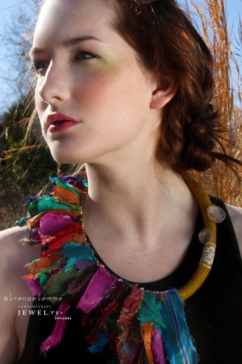 Puede incluir: Una mujer con una blusa negra y un collar de tela colorido y en capas. El collar tiene una base de fieltro amarillo con detalles dorados y una variedad de tiras de tela de colores.