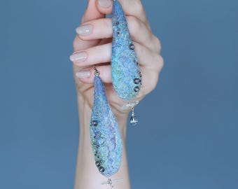 Puede incluir: Un par de pendientes en forma de lágrima hechos de lana de fieltro azul y verde con cuentas plateadas y un dije plateado.