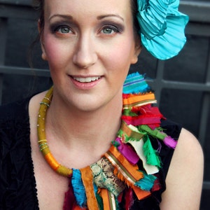 Puede incluir: Una mujer lleva un collar de tela colorido con una flor turquesa en el pelo. El collar est&aacute; hecho de telas de diferentes colores y texturas, y la flor es una flor grande y plisada.