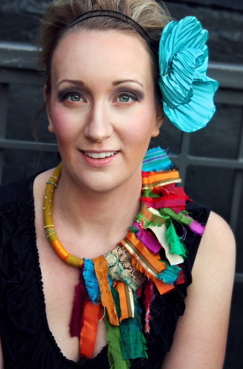 Puede incluir: Una mujer lleva un collar de tela colorido con una flor turquesa en el pelo. El collar est&aacute; hecho de telas de diferentes colores y texturas, y la flor es una flor grande y plisada.