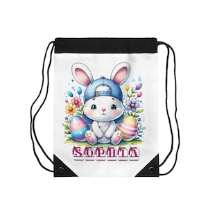 Puede incluir: Bolsa con cordón blanca con un conejito de dibujos animados con una gorra de béisbol azul, rodeado de huevos de Pascua y flores de colores. El nombre "SOPHIA" está impreso en rosa. La bolsa tiene una parte superior negra y cordones negros.