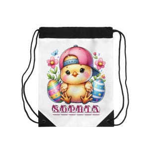 Puede incluir: Bolsa con cordón blanca con un pollito de dibujos animados con una gorra de béisbol rosa, flores y huevos de Pascua. El nombre "SOPHIA" está impreso en la parte inferior. La bolsa tiene cordones negros y una parte superior negra.