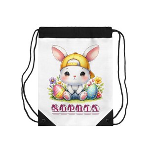 Puede incluir: Bolsa con cordón blanca con un conejito de dibujos animados con una gorra amarilla, rodeado de huevos de Pascua y flores. El nombre "SOPHIA" está impreso en púrpura. La bolsa tiene una parte superior negra y cordones negros.