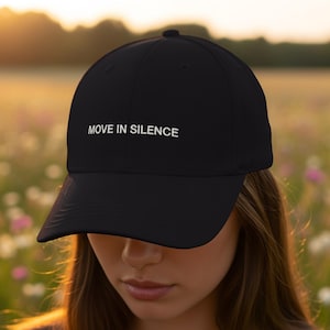 May include: Black baseball cap with the text "MOVE IN SILENCE" in white. The cap is made of a durable material and features a curved brim. The hat is a classic style, suitable for everyday wear.