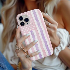 May include: A pastel pink and purple phone case featuring a watercolor American flag design. The case has a light pink background with white stars and vertical stripes in shades of pink and purple. The phone case is held in a person's hands.