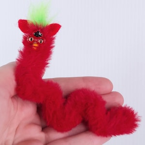 May include: A red plush toy in the shape of a worm, with a Furby-like head. The head has black eyes, orange beak, and gold-colored glasses. The top of the head has a tuft of green feathers. The toy is held in a person's hand.