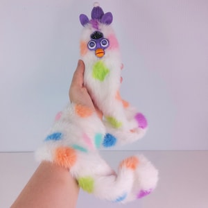 May include: A white plush toy with a long, segmented body and a Furby-like face. The toy features colorful polka dots in orange, pink, blue, green, and purple. The face has purple ears, a knit hat, and a beak. The toy is held in a hand.